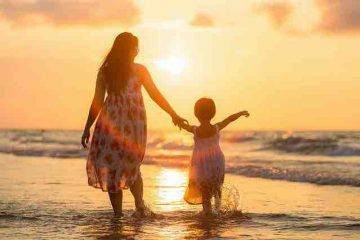A mother holding hands of her daughter and watching the sunrise at a beach depicting mother love, happiness, and care.