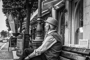 An old man sitting on a bench and looking at the empty road depicting old age, lost hope, solitude, and misery.