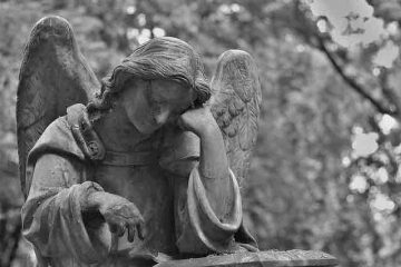 A sad angel in the cemetery kneeling on a grave and thinking depicting past memories of death and misery.
